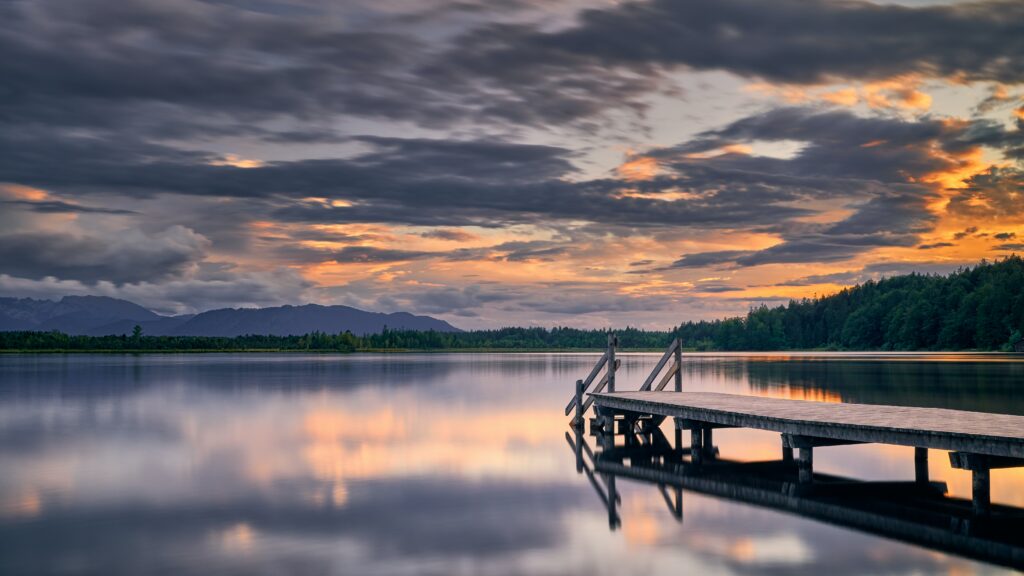 Serene twilight wooden pier over calm lake with colorful sky reflection and lush green trees Wallpaper