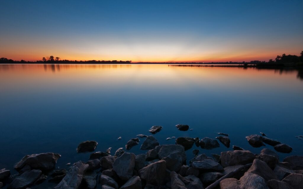 Calm lake at twilight with blue orange sky reflection smooth water and stones foreground wallpaper