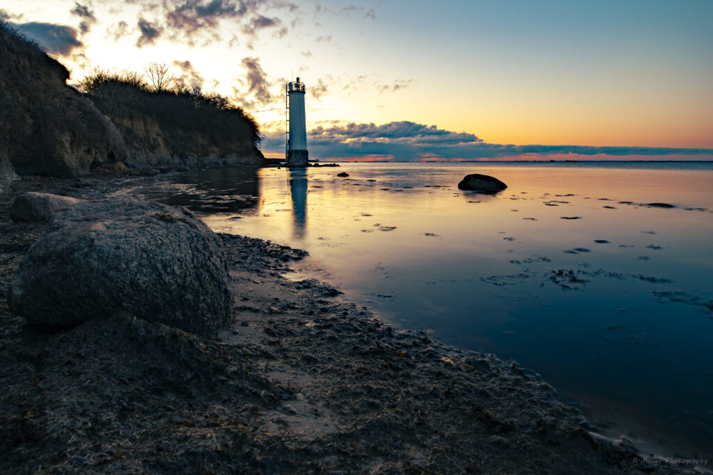 Lone lighthouse silhouette on rocky coast at twilight with vibrant sunset reflecting on calm sea Wallpaper