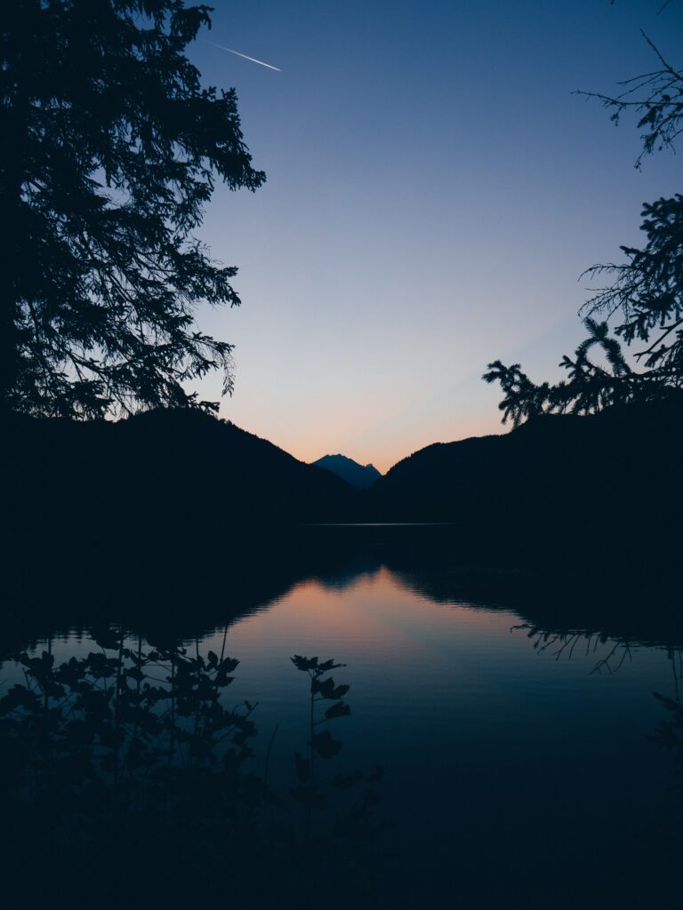 Serene twilight mountain peaks reflected in calm lake with gradient sky and overhanging branches Wallpaper