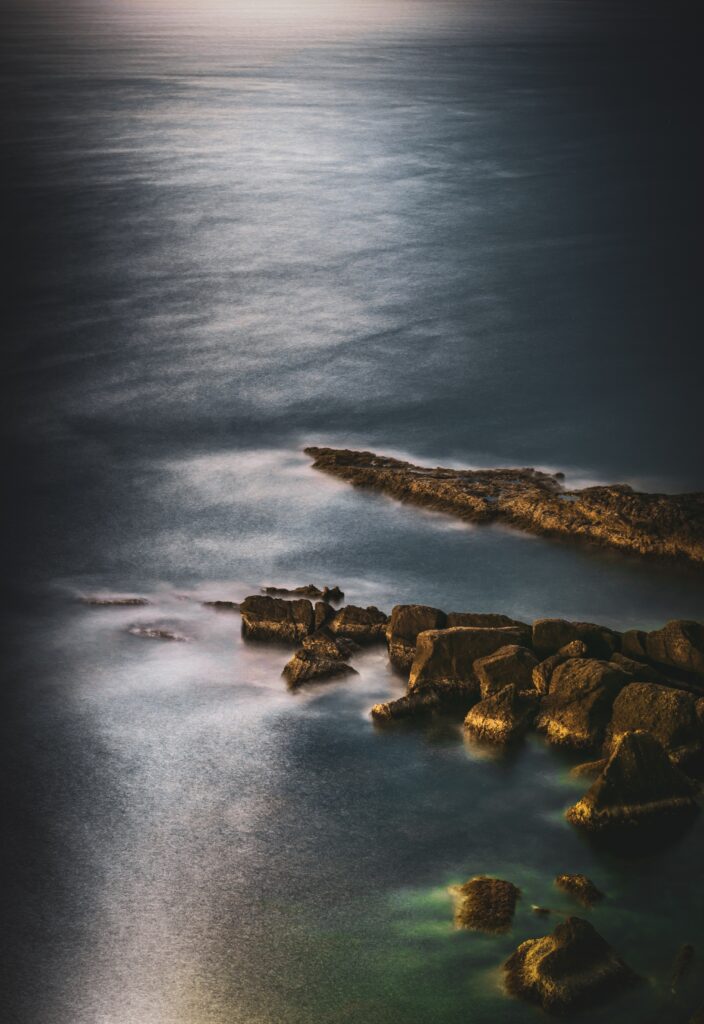 Serene twilight ocean scene with textured stones and soft golden light on gentle blue waves Wallpaper
