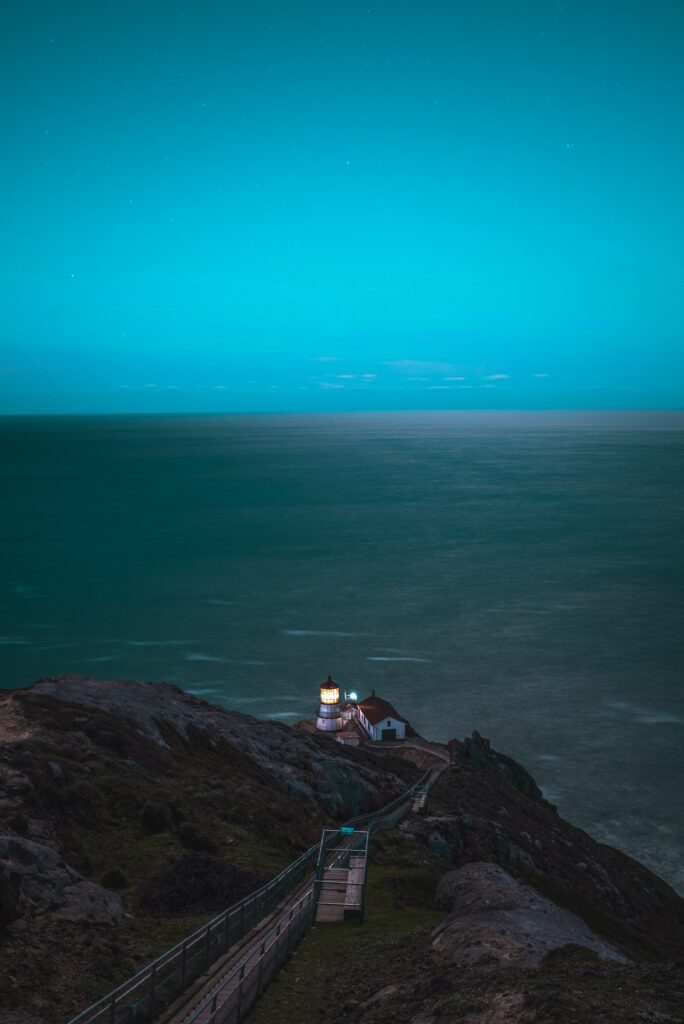 Point Reyes Lighthouse at twilight on rugged cliff overlooking calm ocean with glowing beacon Wallpaper