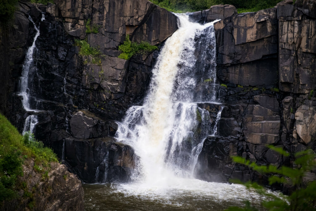 Majestic waterfall cascading dark cliffs with lush green forest and serene stream below nature scene Wallpaper
