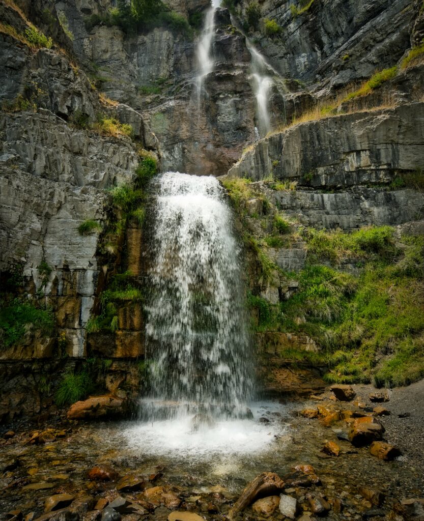 Waterfall cascading over rugged gray rocks surrounded by lush green plants and scattered stones Wallpaper