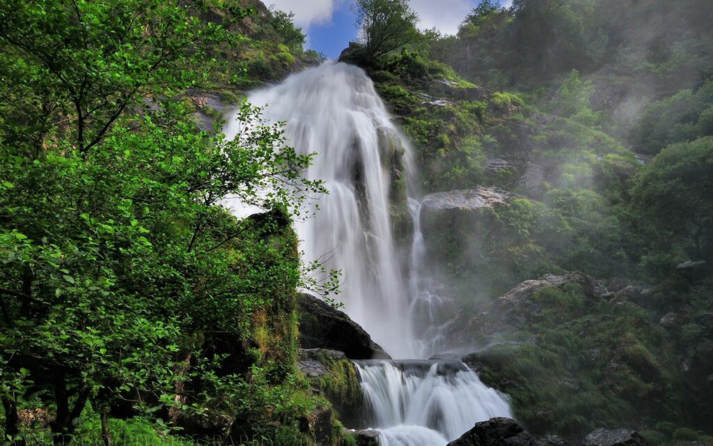 Waterfall cascading over mossy rocks in lush green forest with sunlight and mist background Wallpaper