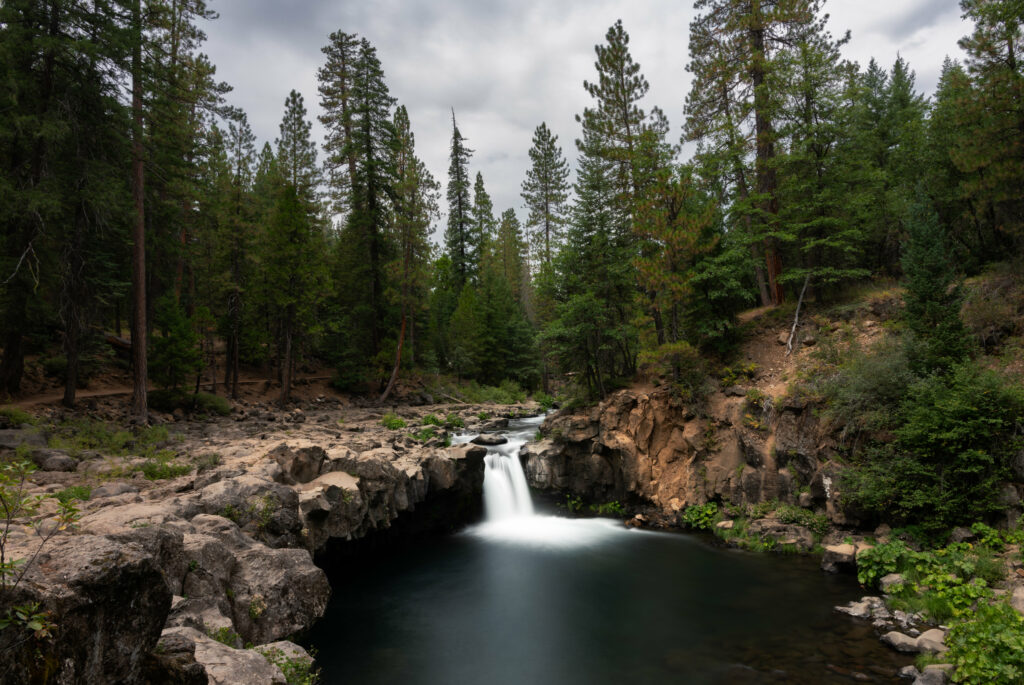 Tranquil waterfall over rocky terrain surrounded by dense green trees under cloudy sky wallpaper