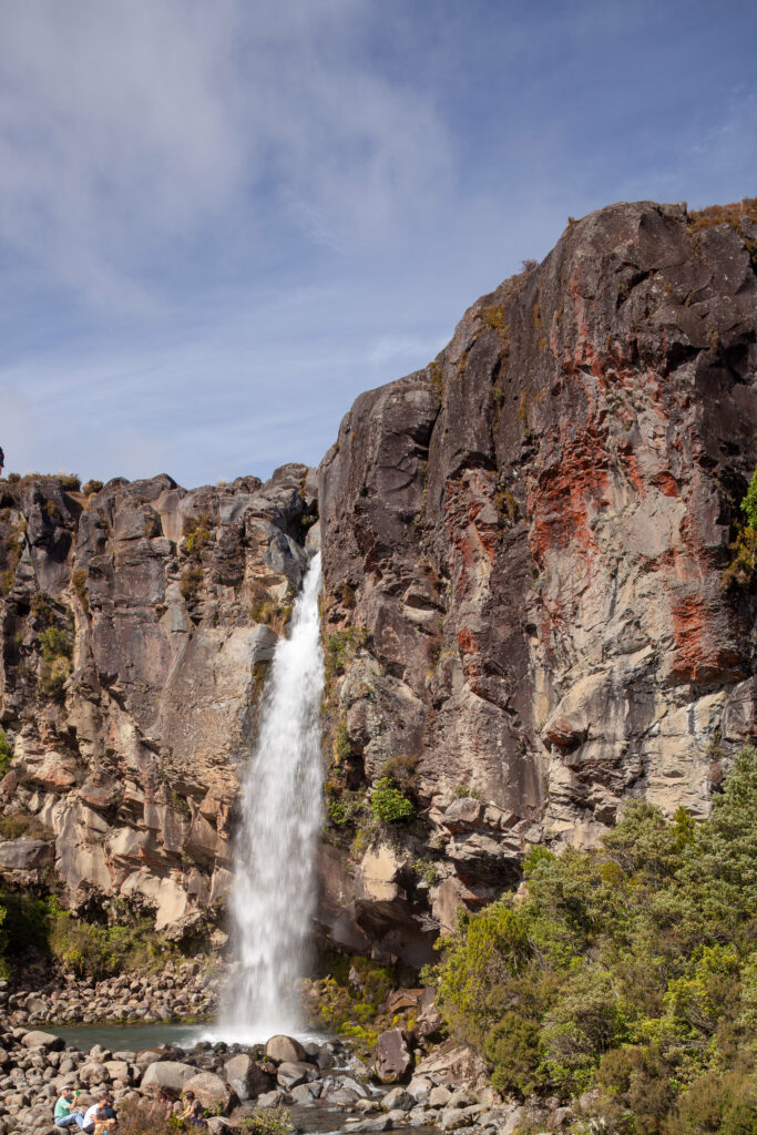 Serene waterfall cascading down rocky cliffs surrounded by lush green trees under blue sky Wallpaper