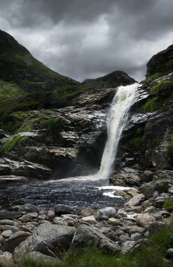 Serene waterfall flowing over rocky cliffs with lush grass under dramatic cloudy sky background Wallpaper