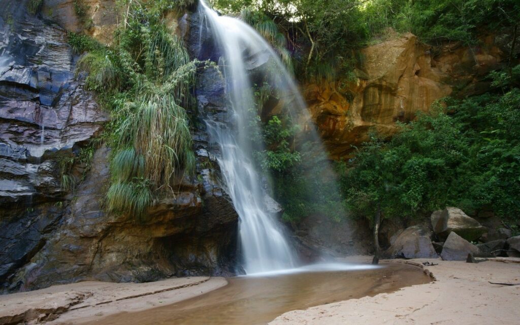 Serene waterfall flowing over rugged rocks into sandy pool surrounded by lush green foliage wallpaper