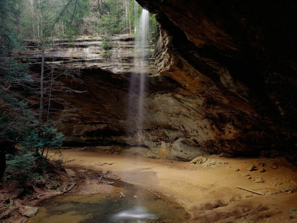 Serene waterfall cascading into sandy grotto surrounded by lush trees and rugged rocks wallpaper