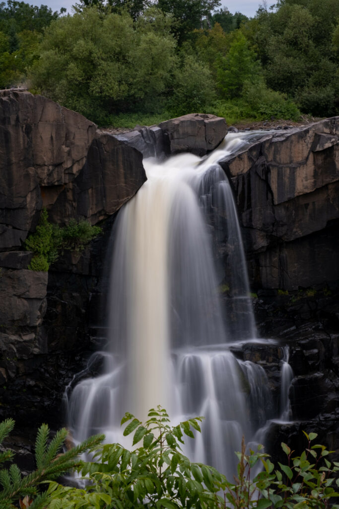 Powerful waterfall cascading over rugged rocks surrounded by lush green foliage and bushes Wallpaper