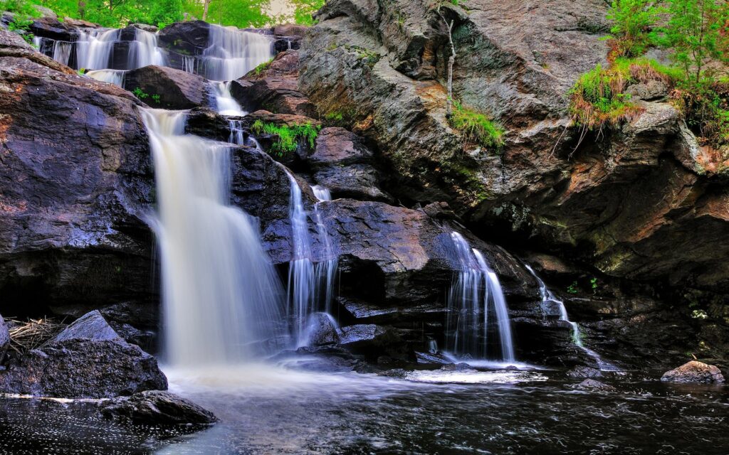 Waterfall cascading over rocks into a river surrounded by lush green foliage vibrant nature scene Wallpaper