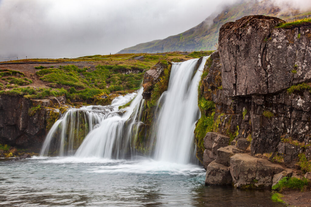 Waterfall cascading over cliffs into a serene pool surrounded by lush greenery and mist Wallpaper