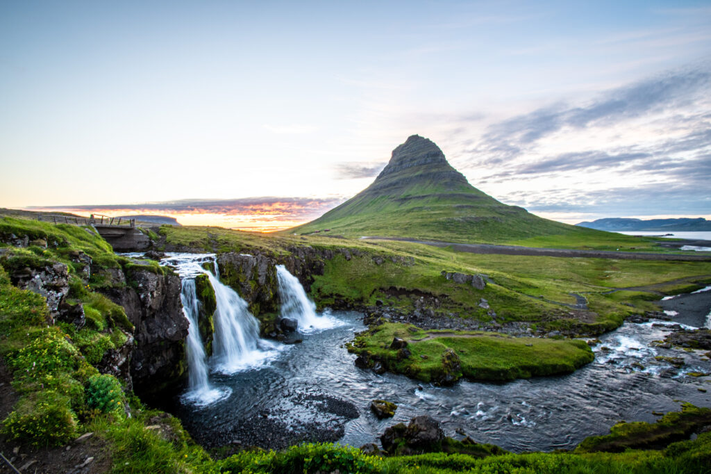 Serene waterfall over mossy stones with lush grass and distant mountain under clear sky wallpaper