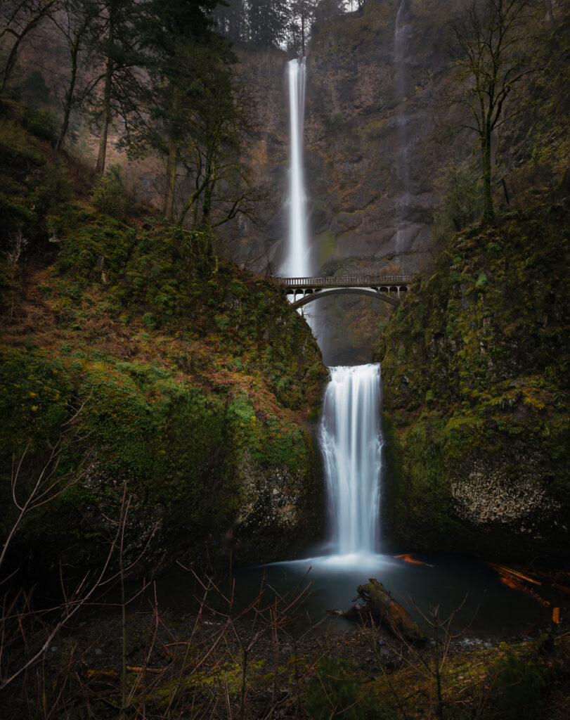 Waterfall cascading over mossy cliffs with arched bridge lush greenery vibrant nature wallpaper