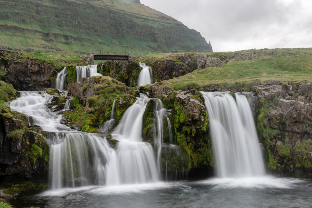 Cascading waterfalls over mossy rocks under wooden bridge with lush mountain and cloudy sky background Wallpaper