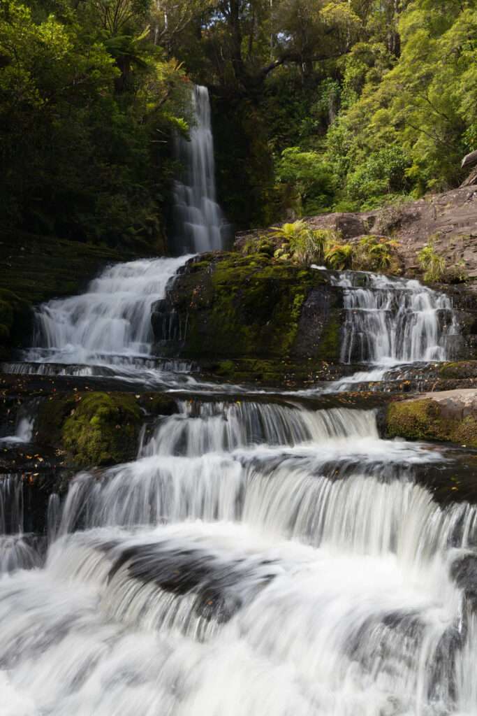Cascading waterfall over mossy rocks in lush green forest with sunlight filtering through trees Wallpaper