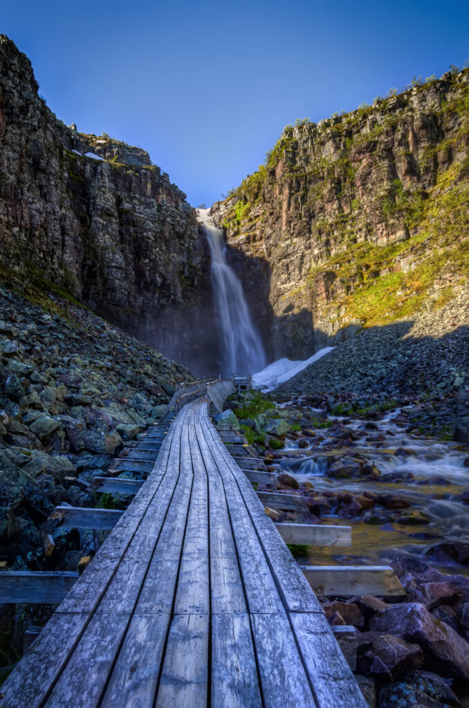 Serene waterfall cascading down rocky cliffs with wooden path under vivid blue sky wallpaper