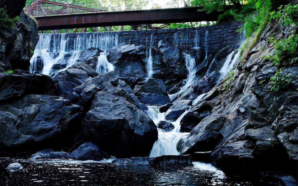 Scenic waterfall over dark rocks with rustic bridge and lush green foliage nature wallpaper background