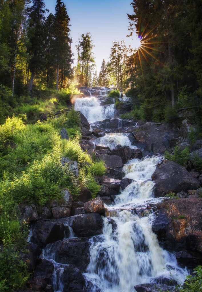 Cascading waterfall over dark stones in lush green forest with sunlight filtering through trees wallpaper