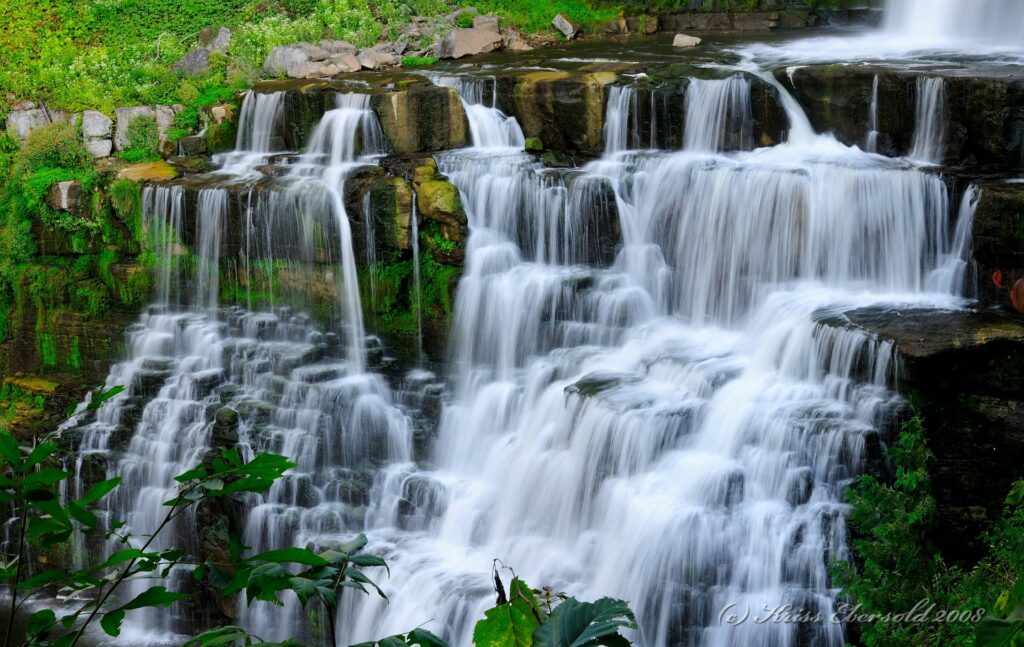 Cascading waterfall over layered rocks with lush green shrubs and vibrant foliage wallpaper background