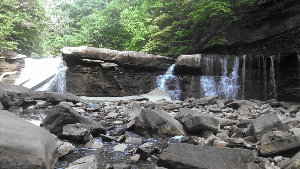 Serene waterfall over layered rocks flowing into river with lush green trees in soft daylight Wallpaper