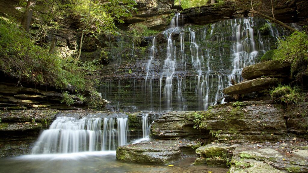 Serene waterfall cascading over layered rocks with mossy wall and lush green foliage background Wallpaper