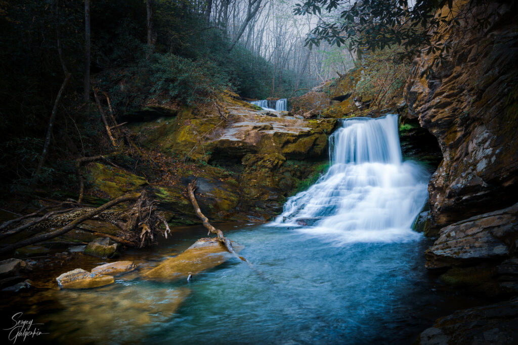 Serene waterfall flowing over mossy rocks surrounded by lush green forest and misty water Wallpaper