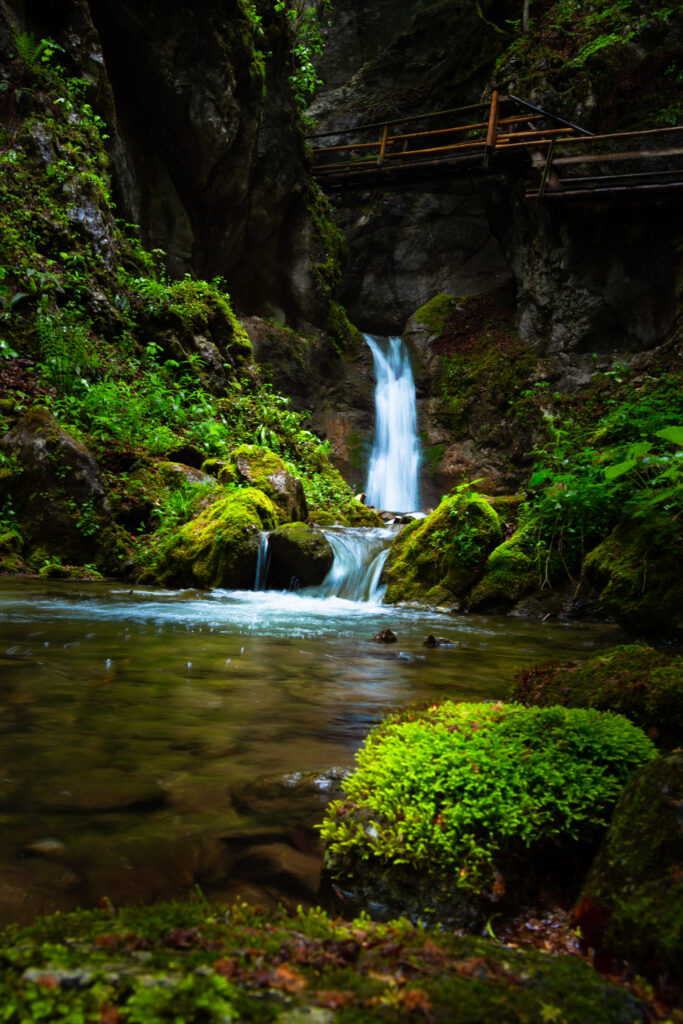 Waterfall cascading over mossy rocks into a tranquil stream with lush greenery and cliffs Wallpaper