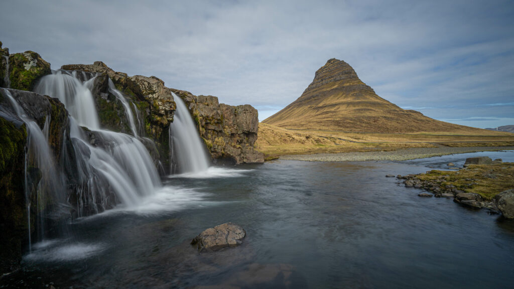 Serene waterfall over mossy rocks with clear river and unique mountain under cloudy sky wallpaper