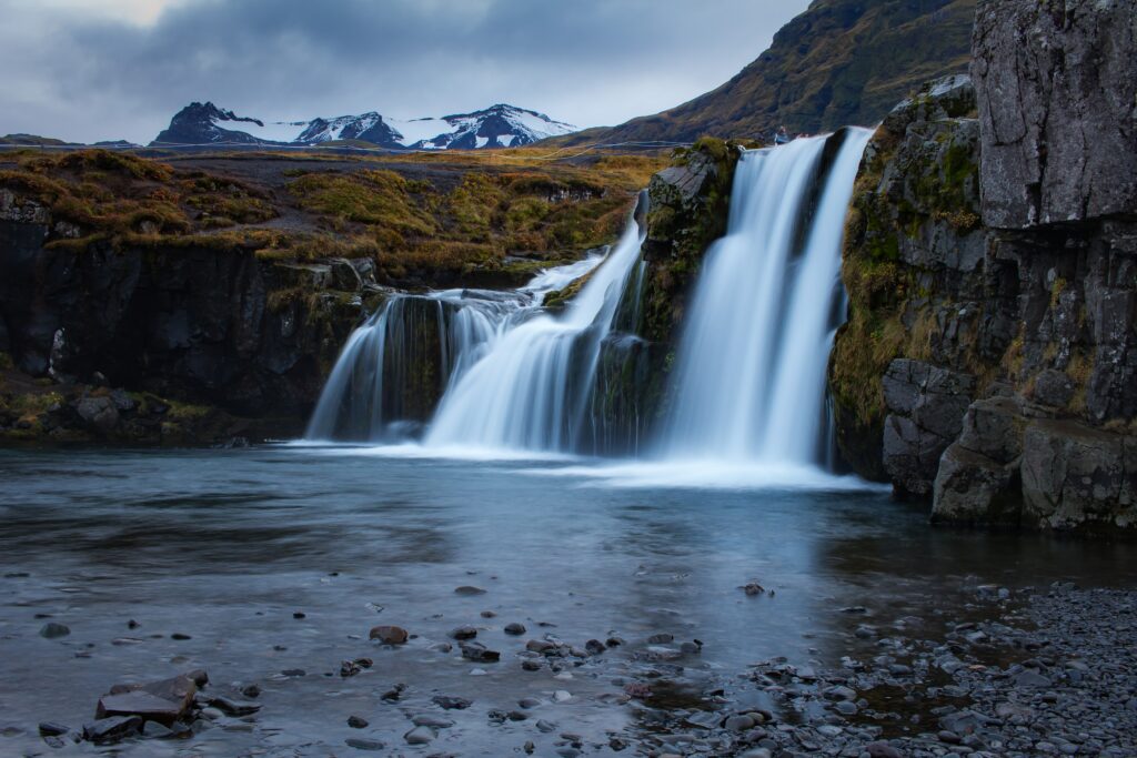 Serene waterfall flowing over mossy rocks with rugged mountains and snowy peaks under clouds Wallpaper