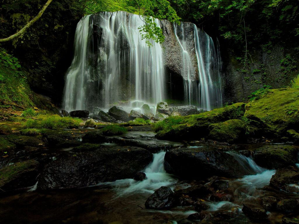 Tranquil waterfall flowing over mossy stones surrounded by vibrant green forest and soft light Wallpaper