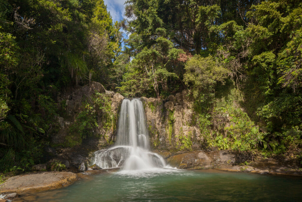 Serene waterfall cascading over rugged rocks with lush green trees and misty spray in daylight Wallpaper