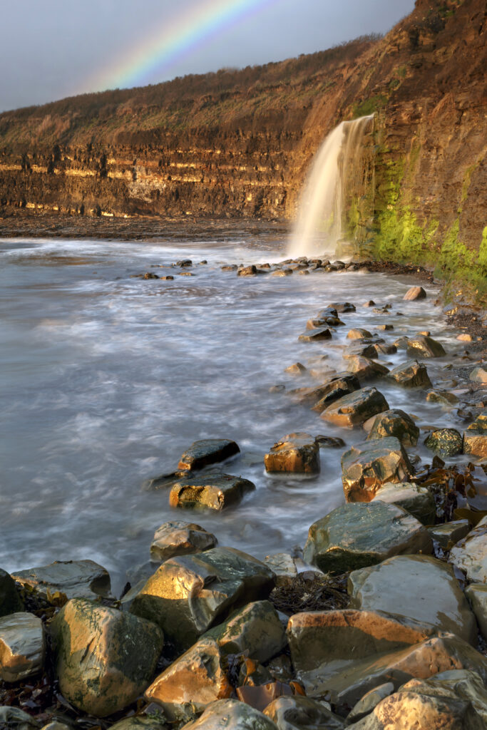 Serene waterfall over cliffs into sea with smooth stones shore and soft rainbow in warm tones Wallpaper