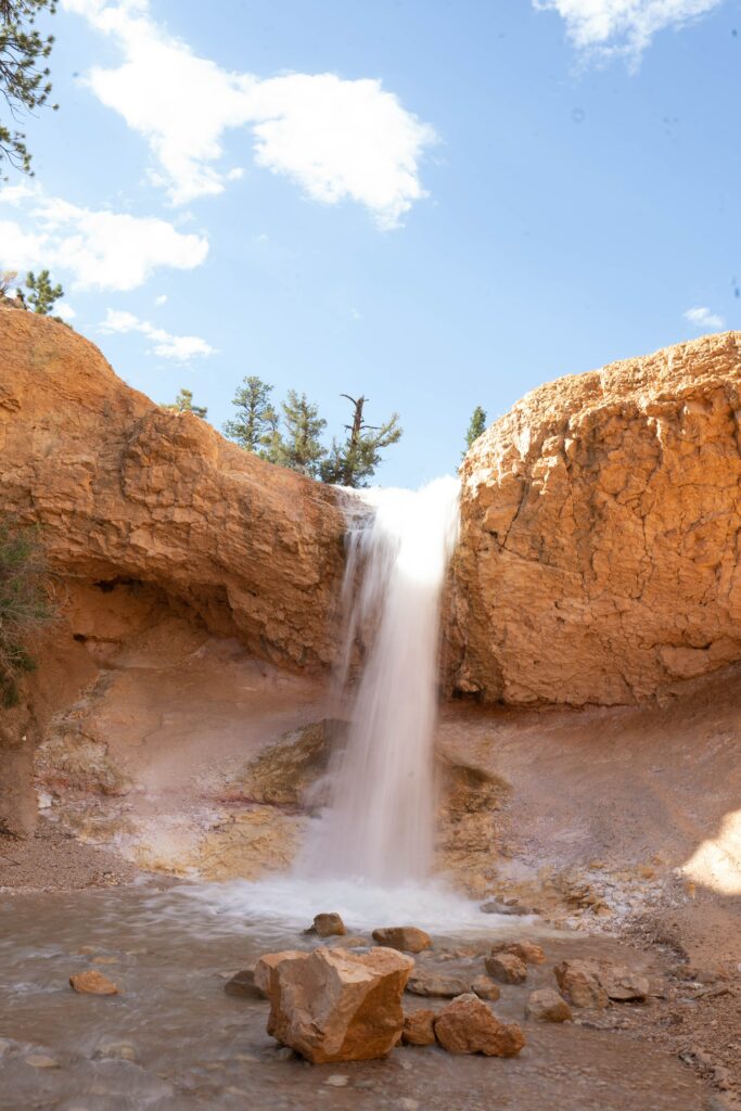 Serene waterfall flowing over rugged cliffs with earthy rocks and clear stream under blue sky Wallpaper
