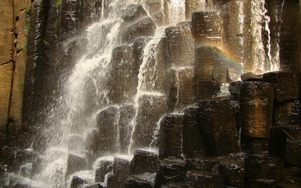 Cascading waterfall over rugged stone formations with sunlight rainbow natural serene scene wallpaper