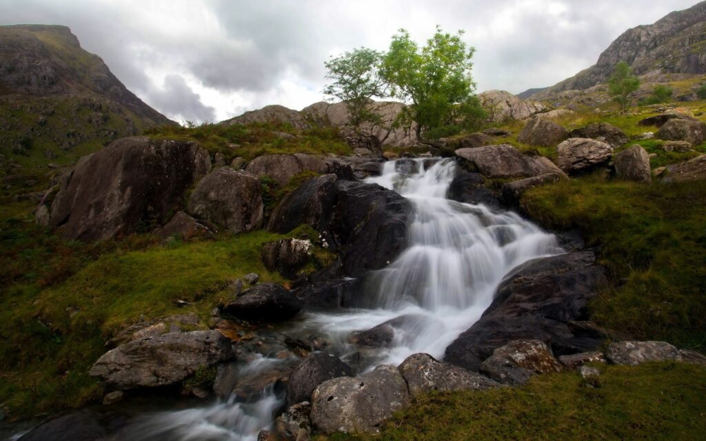 Serene waterfall cascading over rocks with lush grass and dramatic mountain backdrop under clouds Wallpaper