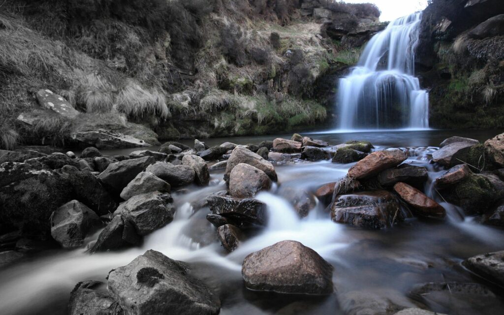 Serene waterfall cascading over rugged rocks into a flowing river with lush green vegetation Wallpaper