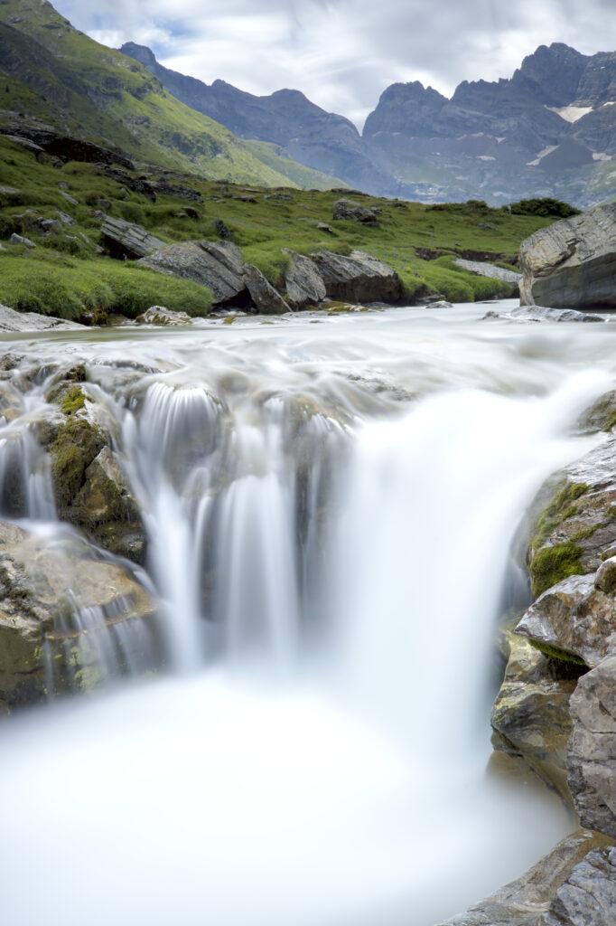 Peaceful waterfall over smooth stones in a lush valley with mountains under a cloudy sky wallpaper