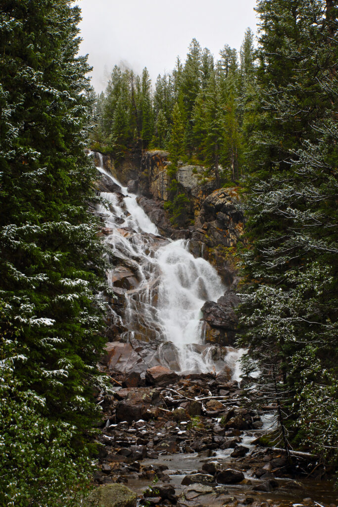 Waterfall cascading over rocky stones surrounded by snow-dusted evergreen trees in winter forest Wallpaper