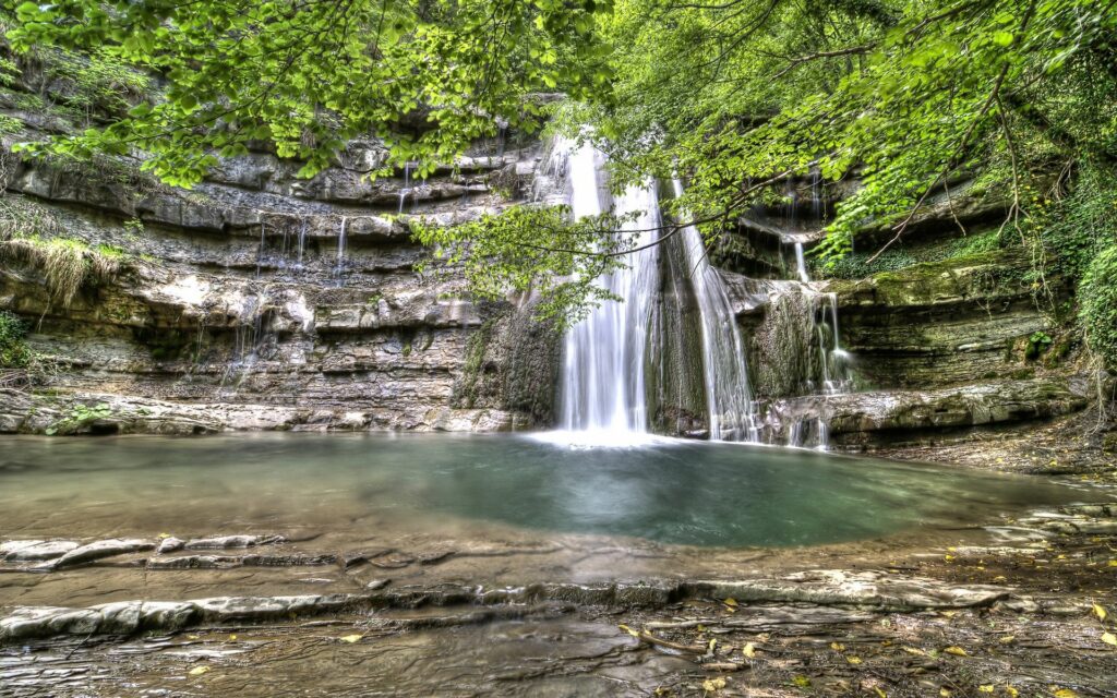 Serene waterfall cascading over rocks into clear pool surrounded by lush green leaves and stones Wallpaper