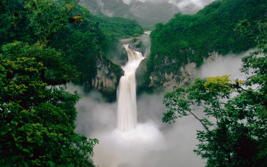 Waterfall cascading into misty gorge surrounded by lush green trees serene nature wallpaper background