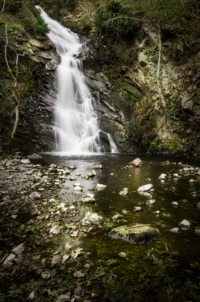 Serene waterfall cascading over dark stones into a tranquil stream with soft green earthy tones Wallpaper