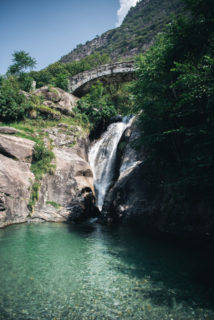 Serene waterfall over smooth stones into clear lake with stone bridge lush greenery and hills background Wallpaper