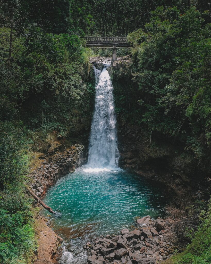 Serene waterfall over clear blue lake surrounded by green trees rocky terrain and stone bridge Wallpaper
