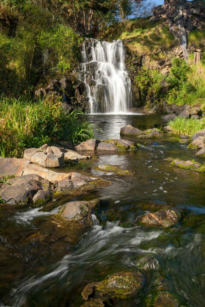 Peaceful waterfall over rocks with clear stream and lush green foliage in sunlight wallpaper background