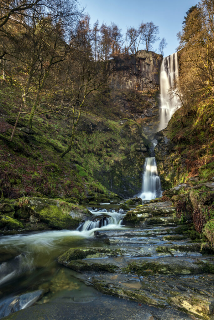 Serene waterfall over mossy rocks with lush trees under clear blue sky nature background wallpaper