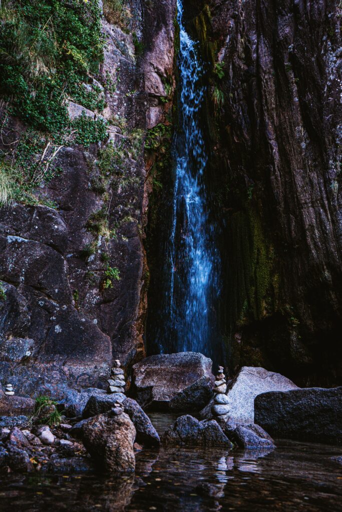 Serene waterfall flowing over rocky cliffs with balanced stone stacks in cool earth tones background Wallpaper