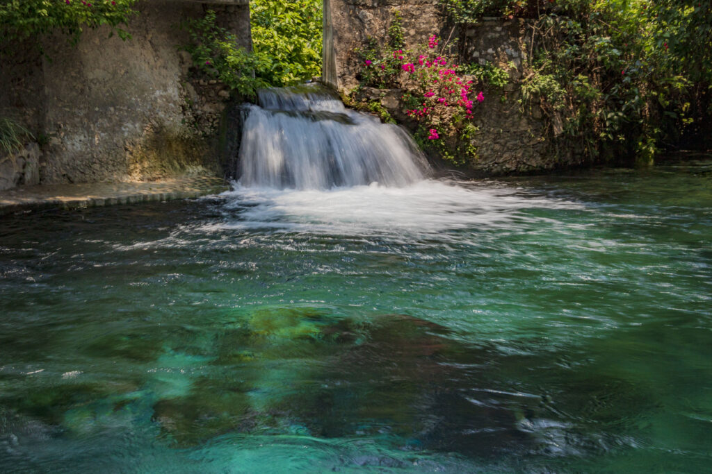 Small waterfall flowing into turquoise water surrounded by green trees and vibrant pink flowers Wallpaper