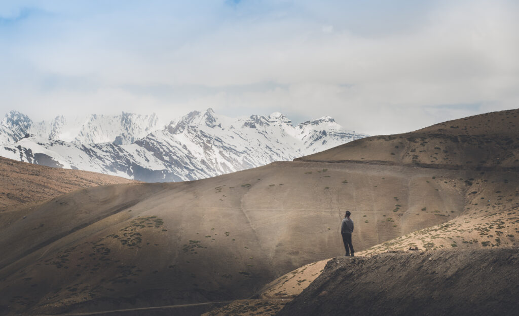 Solitary man amid rugged hills gazing at snow-capped mountains under cloudy sky wallpaper